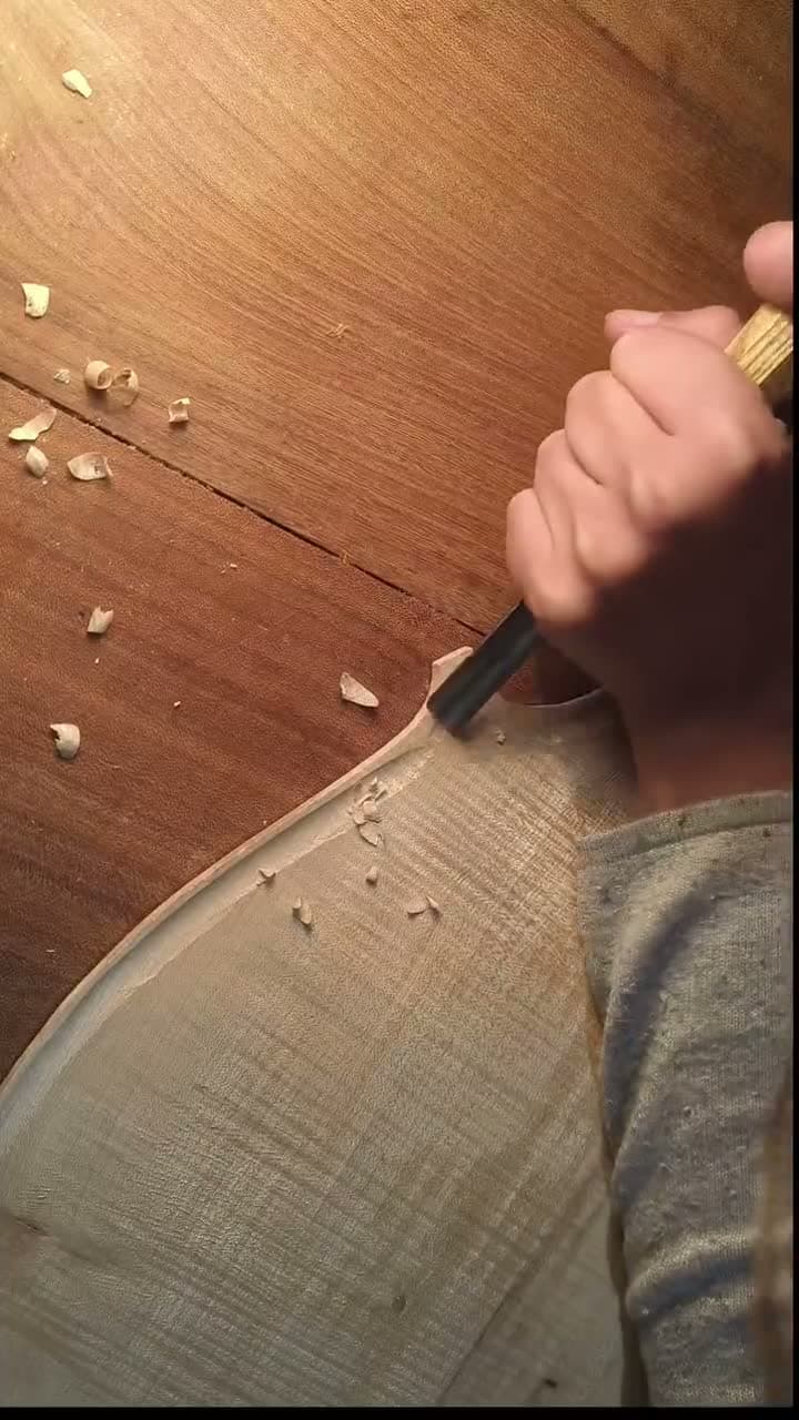 Main tenant une gouge, en train de creuser la table d’un violoncelle, copeaux de bois autour