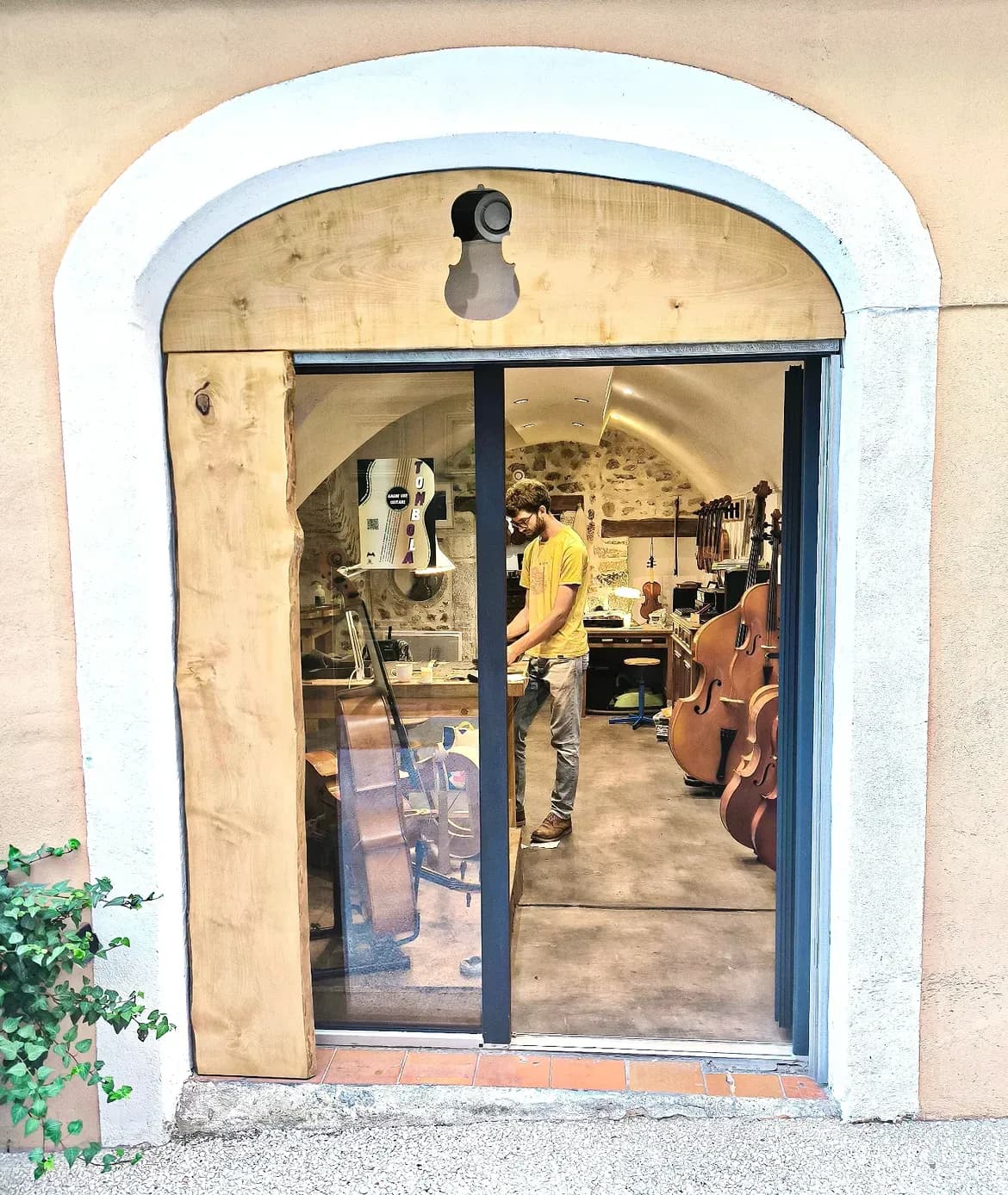 La porte vitrée de l’atelier ouverte sur la rue, un luthier au travail et des violoncelles suspendus au mur du fond