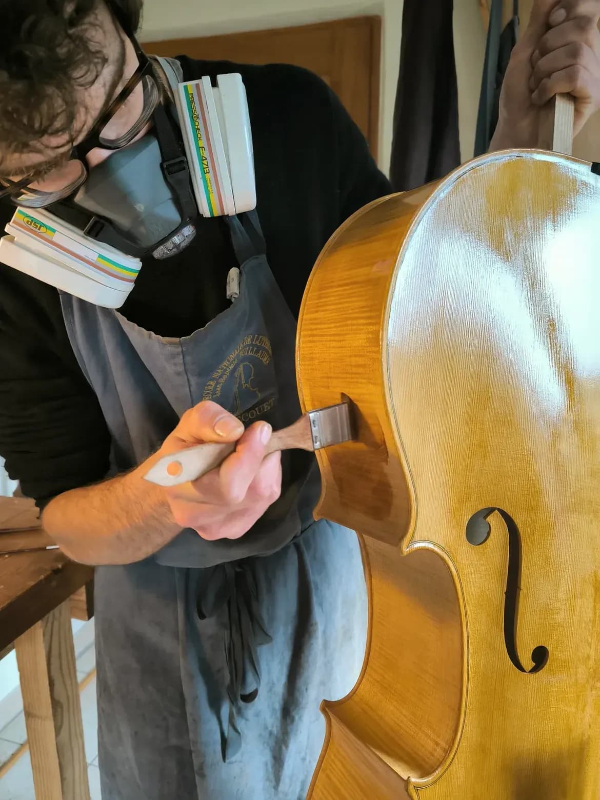 Un luthier masqué applique au pinceau la première couche de vernis sur la table d’un violoncelle, dont la volute est tenue par une seconde personne